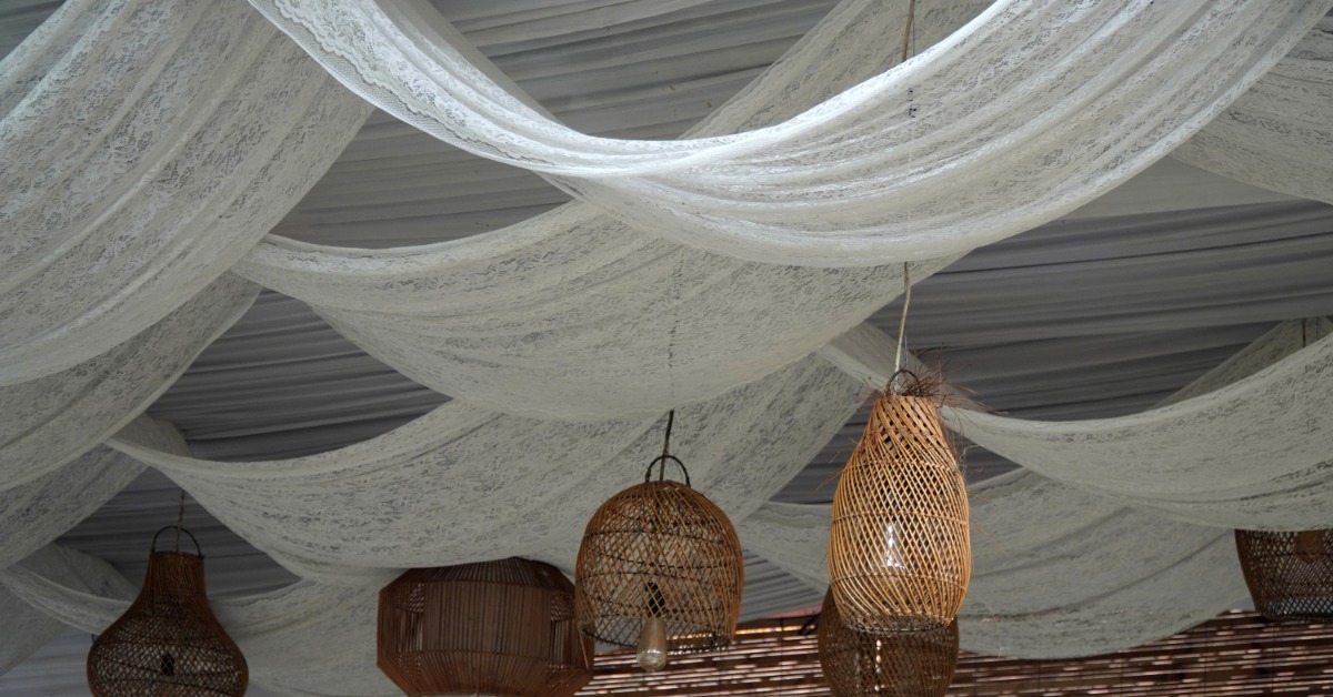 Delicate white ceiling drapery flowing across the ceiling in a large room. Six rattan lamps hang from the ceiling.